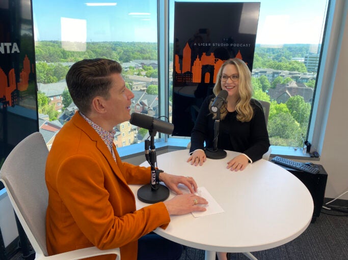 Man in orange jacket interviewing blonde woman with glasses
