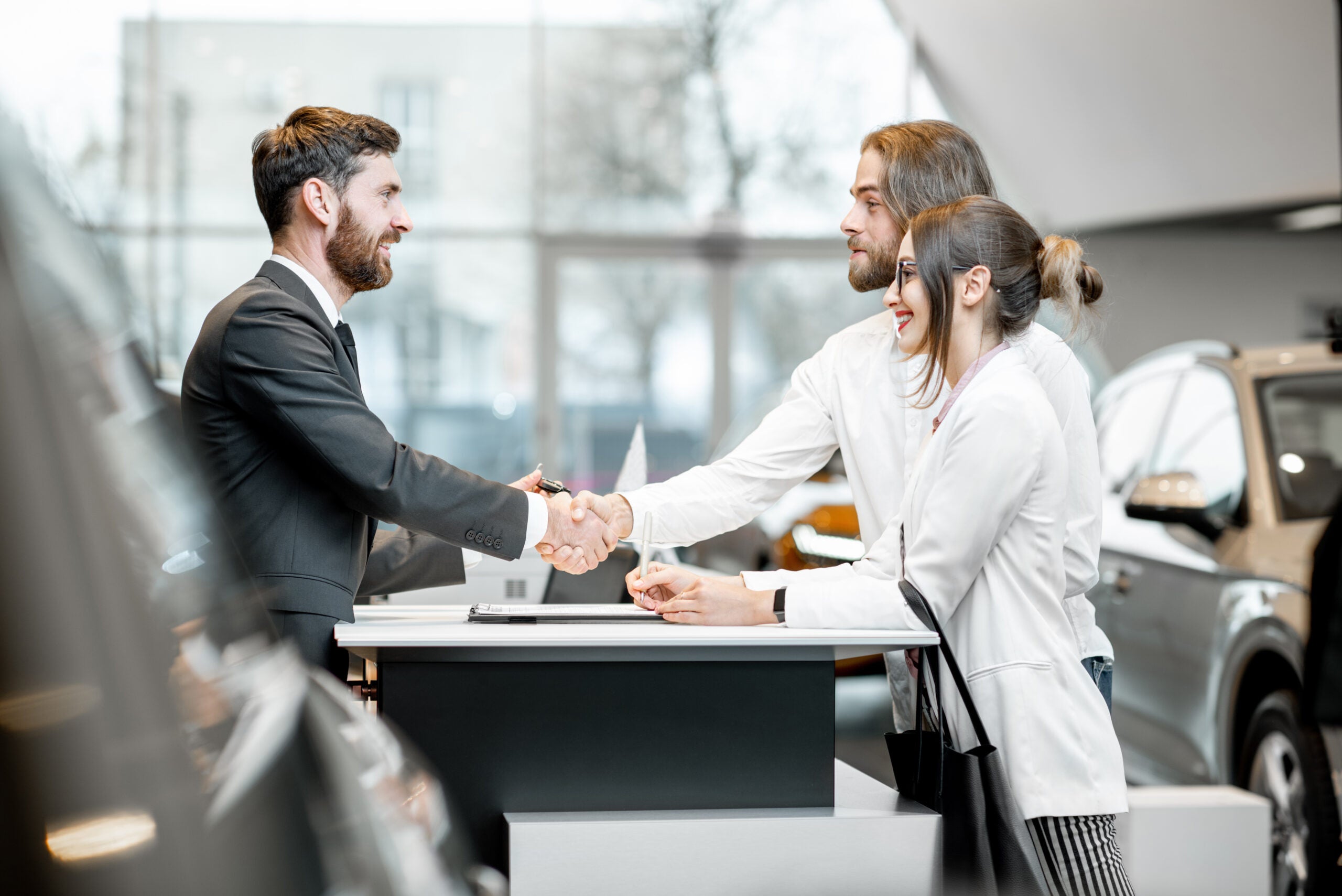 Young business couple shaking hands having a deal with salesperson at the stand buying or renting car in the showroom