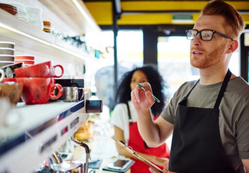Pensive male waiter in apron counting cups standing at bar with female partner during working process.Multicultural employees of coffee shop making inventory to do report