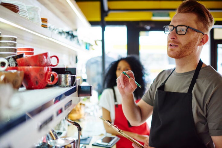 Pensive male waiter in apron counting cups standing at bar with female partner during working process.Multicultural employees of coffee shop making inventory to do report