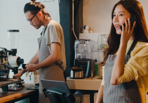 Young female barista taking order by mobile phone and digital tablet standing behind bar counter while talking with customer on mobile phone at cafe restaurant. Owner small business concept.
