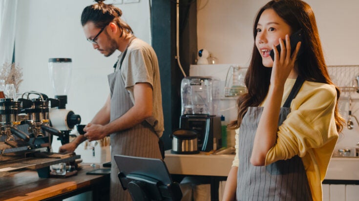 Young female barista taking order by mobile phone and digital tablet standing behind bar counter while talking with customer on mobile phone at cafe restaurant. Owner small business concept.