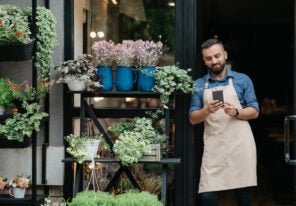Social media, ad and online offer for small business owner. Smiling bearded millennial european male in apron watch on smartphone at front door of flowers rustic shop with different plants, free space