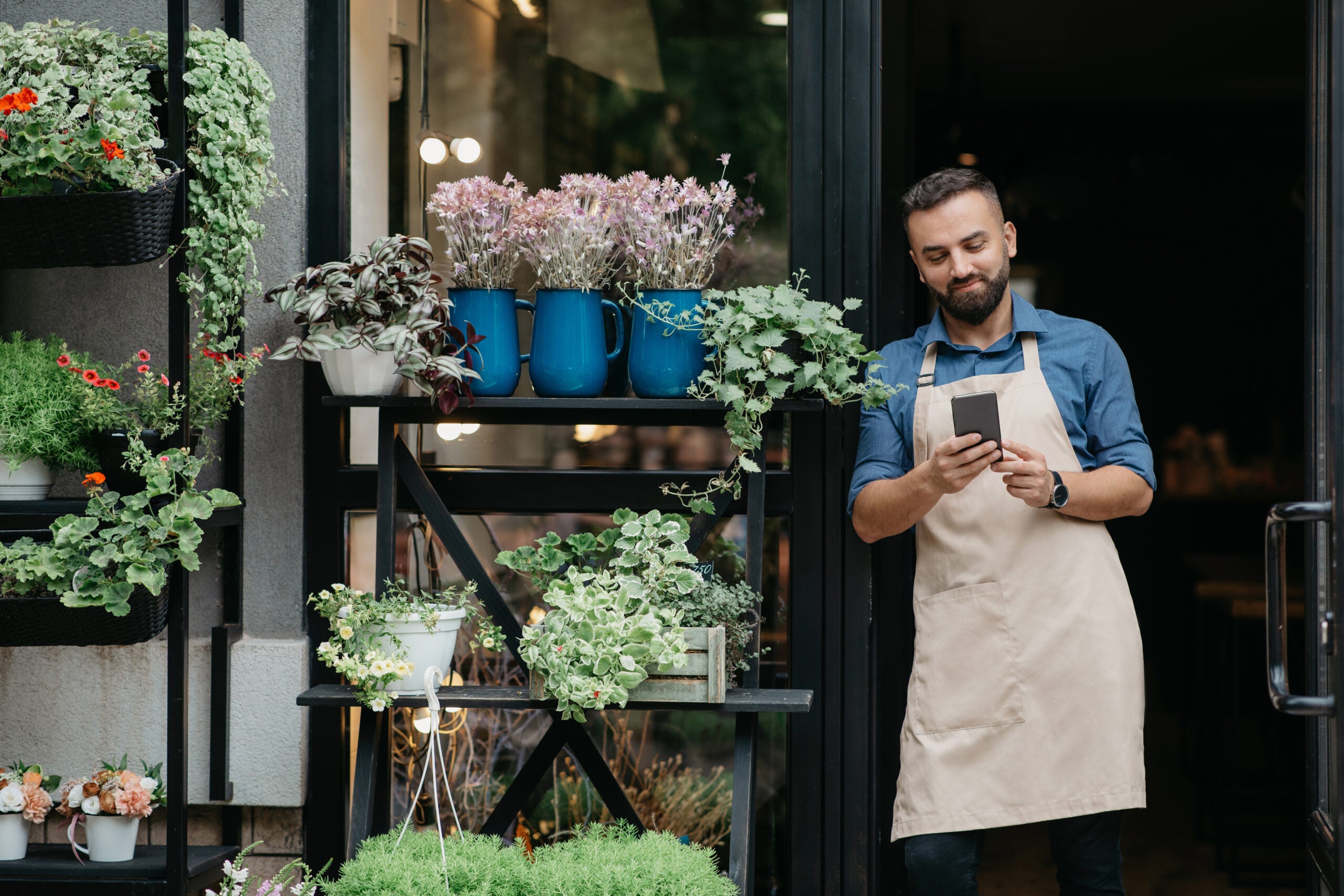 Social media, ad and online offer for small business owner. Smiling bearded millennial european male in apron watch on smartphone at front door of flowers rustic shop with different plants, free space