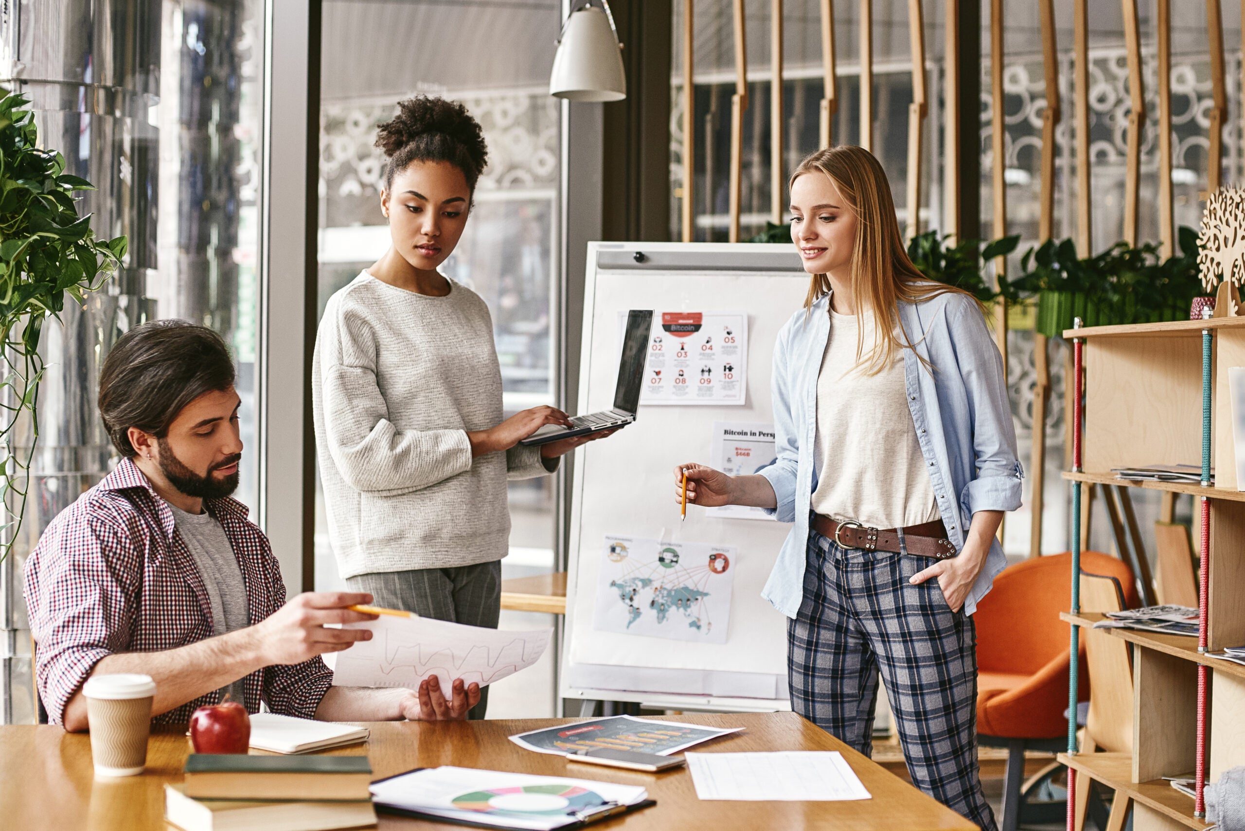 Attractive young woman standing near whiteboard with a laptop in her hands and looking to her male colleague, that holds a chart with the results. Another colleague looks at it with a smile. Concept of success