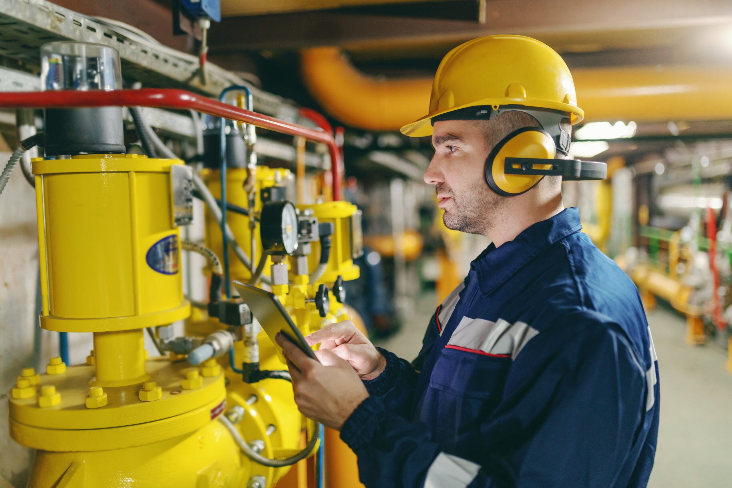 Profile of hardworking worker with helmet, antifones and in protective suit checking air pressure on boilers while standing in heavy industry plant