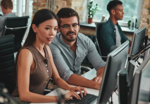 Two people looking at computer monitor and discussing work tasks while their colleagues working on the background.