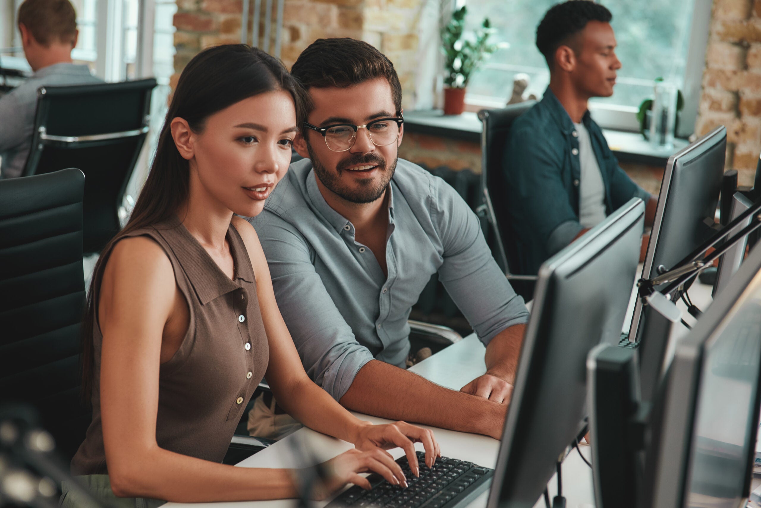 Two people looking at computer monitor and discussing work tasks while their colleagues working on the background.