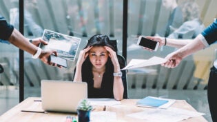 Young disappointed woman feeling headache from deadline surrounded by colleagues at workplace, multi-purpose female employee,tired of work and exhausted, stress in office. Overworked female in confuse