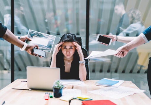 Young disappointed woman feeling headache from deadline surrounded by colleagues at workplace, multi-purpose female employee,tired of work and exhausted, stress in office. Overworked female in confuse