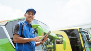 a bus crew member in uniform and a cap smiling at the camera while using a digital tablet against the background of the bus fleet