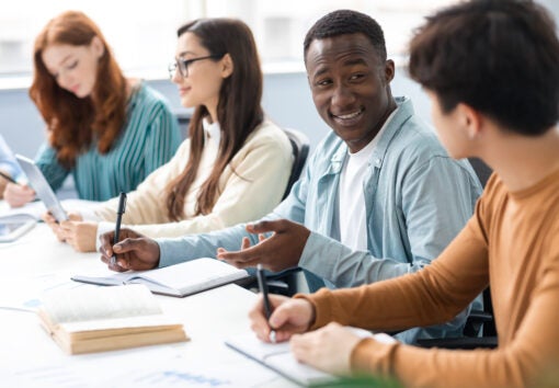 Cheerful colleagues enjoying working process and cooperation, writing in notebooks and talking, skilled students preparing homework task together doing group project in college library