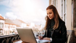 Young bussines woman working outdoors. Writting in a notebook while sitting in front of a laptop.