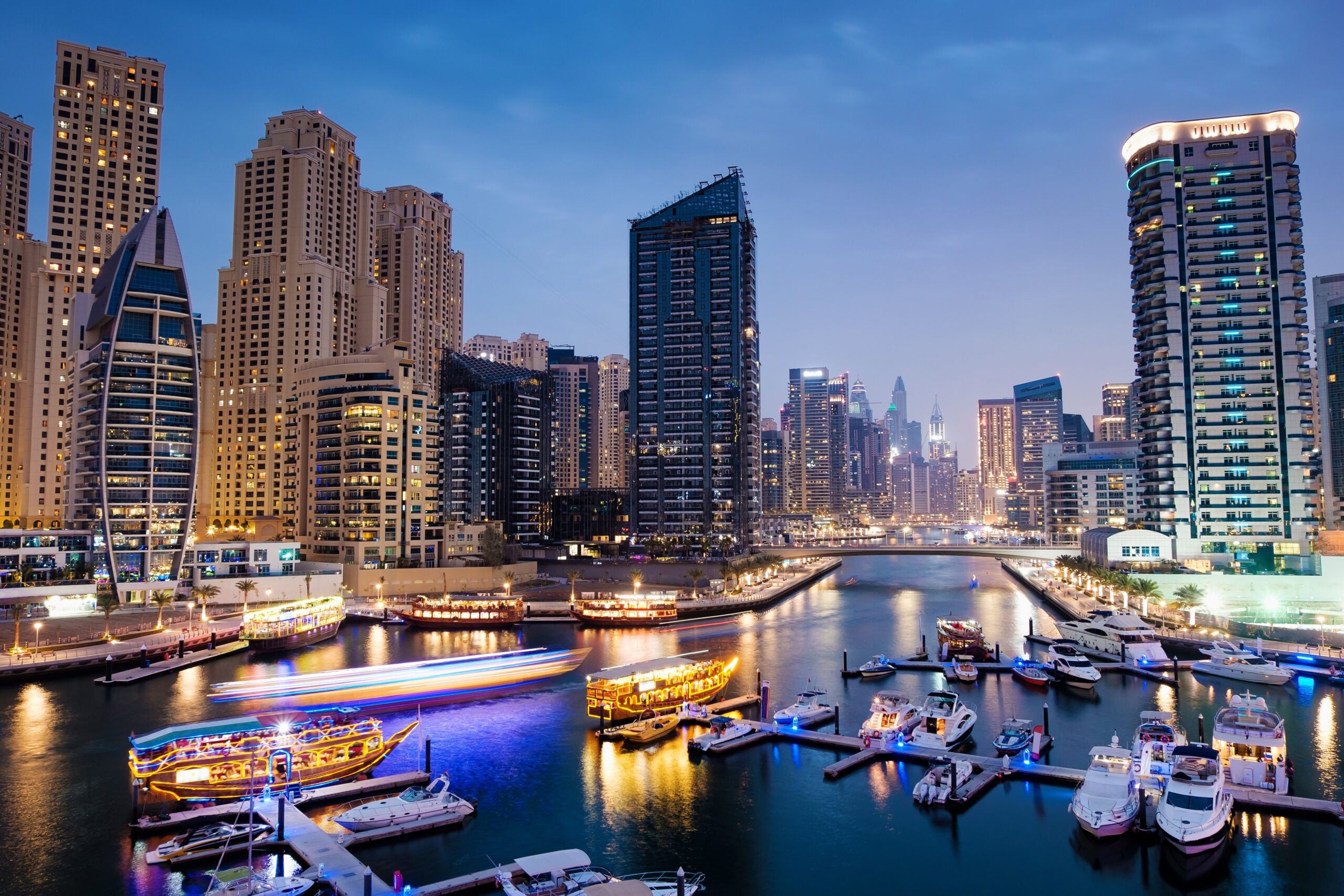 Dubai marina with boats and buildings with gates at night with lights and blue sky, United Arab Emirates