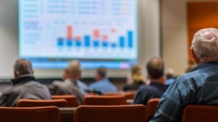 A senior citizen sits in a lecture hall listening to a speaker present a financial planning seminar