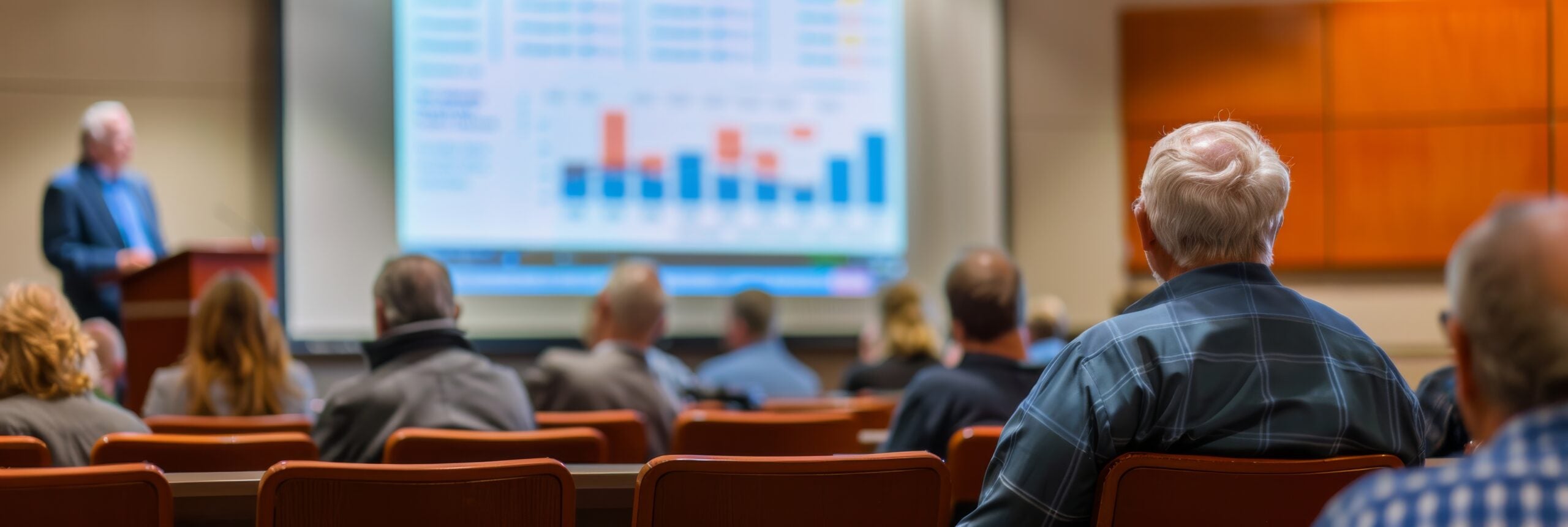 A senior citizen sits in a lecture hall listening to a speaker present a financial planning seminar