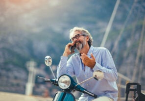 Mature man on motor scooter using phone in marina.