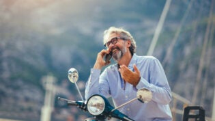 Mature man on motor scooter using phone in marina.