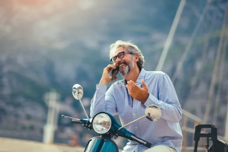 Mature man on motor scooter using phone in marina.