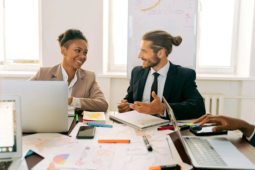 Woman and man in business attire smiling at each other over desk filled with paperwork