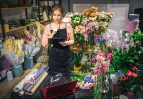 young florist hold black planchette. She think and look at it. Florist is surround with flowers and plants.