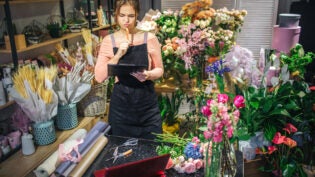 young florist hold black planchette. She think and look at it. Florist is surround with flowers and plants.