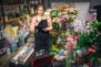 young florist hold black planchette. She think and look at it. Florist is surround with flowers and plants.