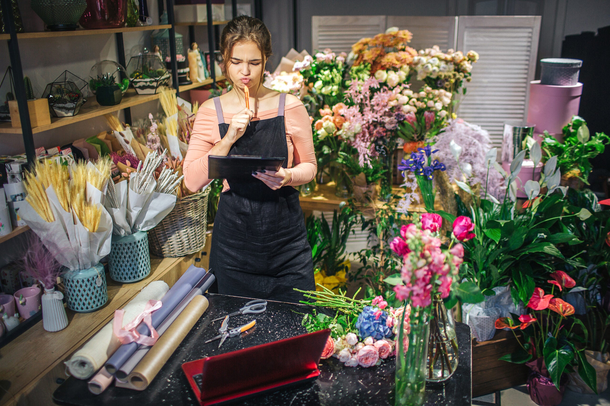 young florist hold black planchette. She think and look at it. Florist is surround with flowers and plants.