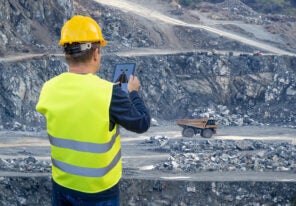 An engineer holds a tablet in his hand and remotely controls a heavy-duty dump truck in the background of a mining quarry