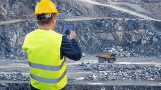 An engineer holds a tablet in his hand and remotely controls a heavy-duty dump truck in the background of a mining quarry