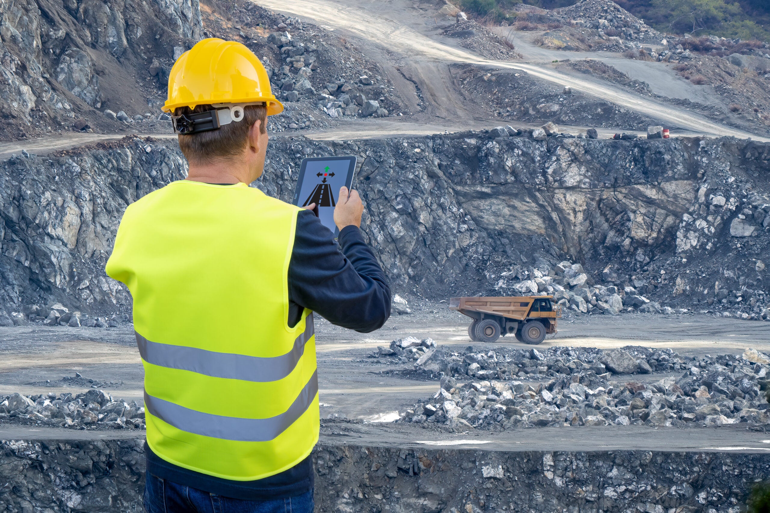 An engineer holds a tablet in his hand and remotely controls a heavy-duty dump truck in the background of a mining quarry