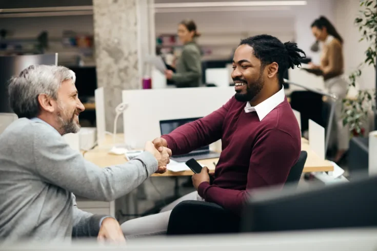 Happy male entrepreneurs shaking hands while greeting at corporate office.