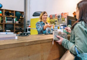 Female customer giving credit card to smiling woman shop assistant to pay purchase on local store