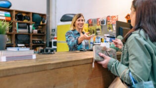 Female customer giving credit card to smiling woman shop assistant to pay purchase on local store