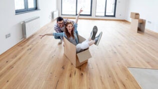 Young couple in new empty room of house. She is sitting on card box while he pushing her from behind