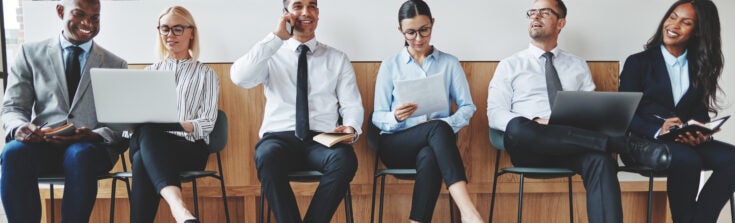 Group of diverse businesspeople smiling while sitting in a row in an office reception waiting for a meeting