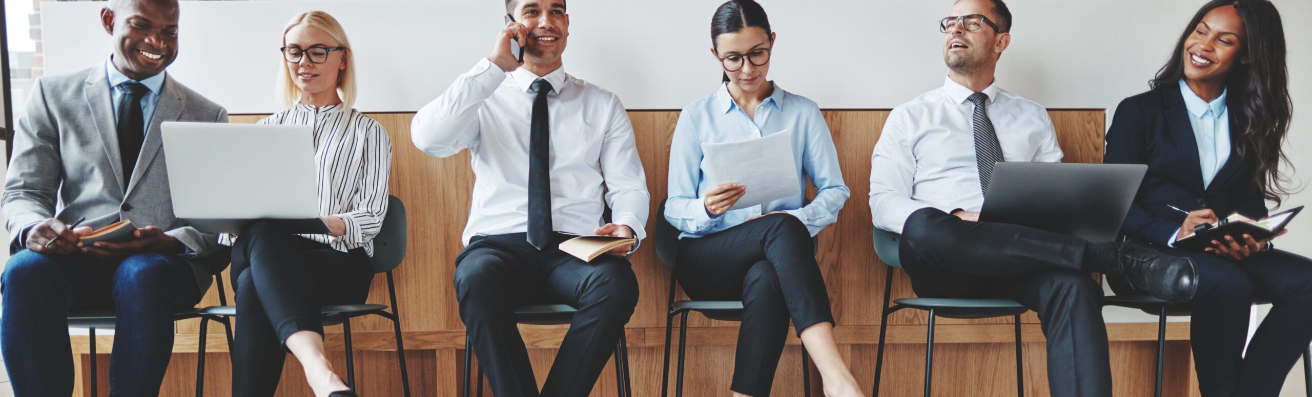 Smiling diverse businesspeople sitting together in an office rec Group of diverse businesspeople smiling while sitting in a row in an office reception waiting for a meeting