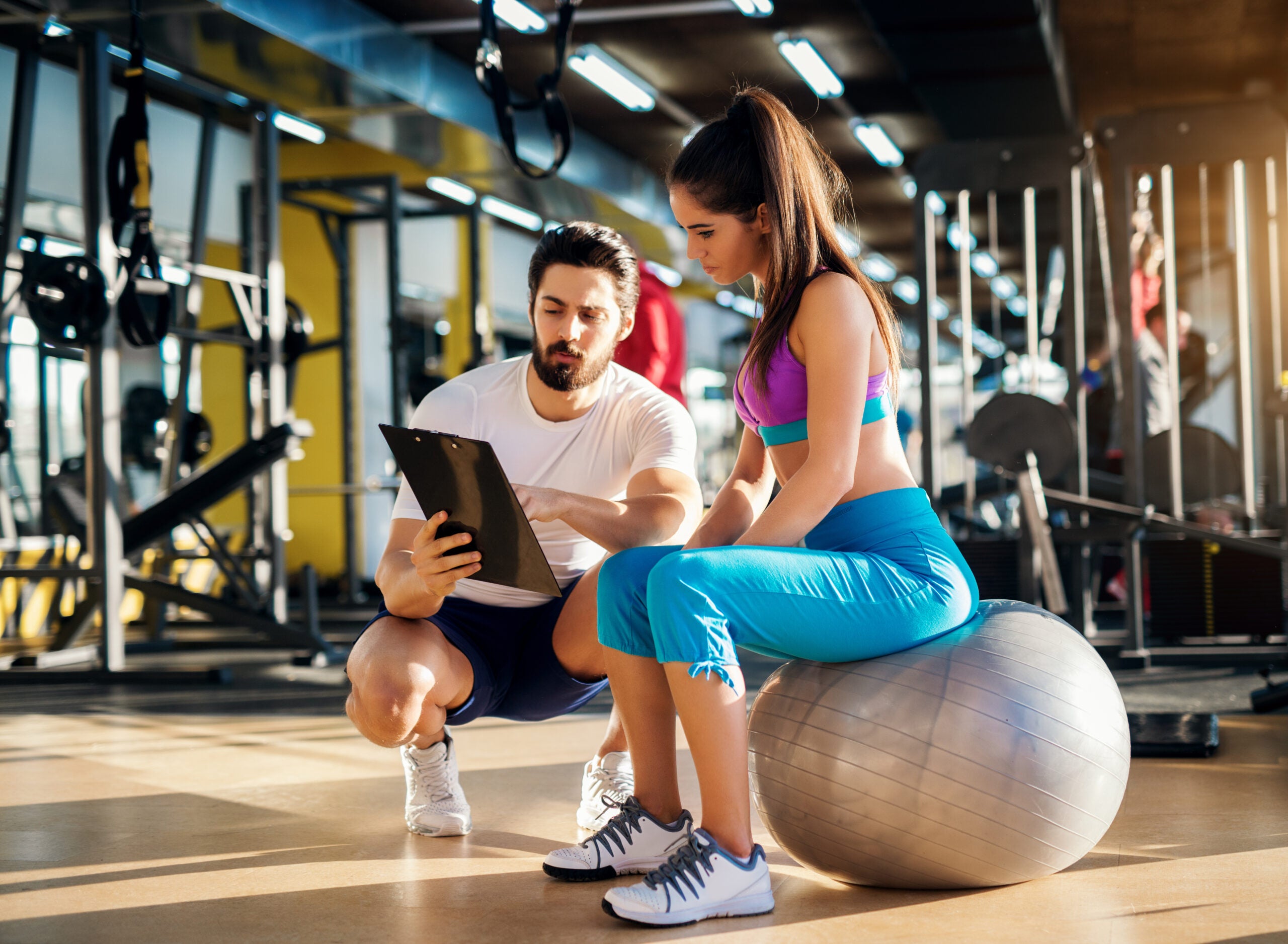 Young healthy active woman sitting on the gym ball and consulting with a personal trainer about an exercise plan. Young healthy active woman sitting on the gym ball and consulting with a personal trainer about an exercise plan.