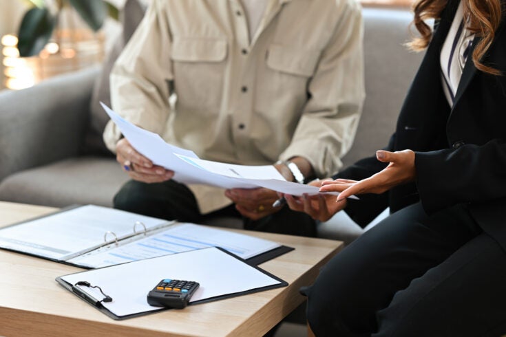 Close up male senior client and female insurance agent sitting on a couch in a house pointing out the agreement condition together, for real estate, business, finance and insurance concept.